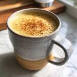 Close-up of a steaming mug filled with Christmas Hot Buttered Rum, topped with frothy foam and a sprinkle of cinnamon.