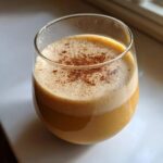 A festive glass of Christmas Spiked Chai Eggnog, topped with foam and a sprinkle of cinnamon.