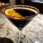 A spooky Halloween Black Magic Martini in a glass, rimmed with sugar and garnished with an orange slice.