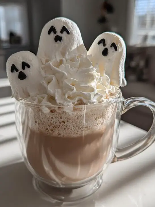 A clear mug filled with Halloween Ghostly White Hot Chocolate, topped with whipped cream and three marshmallow ghosts.