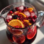 A clear glass pitcher filled with Halloween Hocus Pocus Sangria, featuring oranges, cranberries, blackberries, pomegranate seeds, and ice.