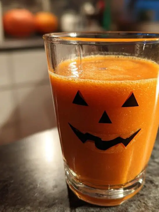 A vibrant orange Halloween Jack-O’-Lantern Smoothie fills a glass decorated with a jack-o'-lantern face.