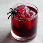 A spooky Halloween Vampire's Kiss Cocktail in a glass, garnished with a black spider and filled with ice and deep red liquid.