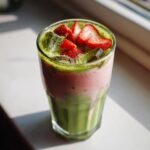 A tall glass of Iced Strawberry Matcha Latte with layers of green matcha, pink strawberry milk, ice, and fresh strawberry slices.