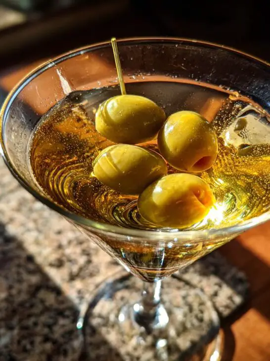 A close-up of a New Year’s Eve Black Tie Martini garnished with four green olives on a toothpick.