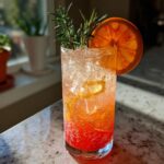 A tall glass filled with New Year’s Eve Citrus Sparkler Mocktail, ice, orange slices, and rosemary garnish.