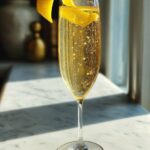 A tall glass filled with bubbly New Year’s Eve Elderflower Champagne Fizz, garnished with a lemon twist.