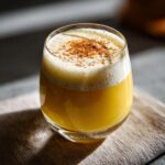 A festive glass of Thanksgiving Bourbon Vanilla Eggnog, topped with foam and a sprinkle of cinnamon.