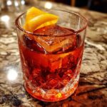 A close-up of a Thanksgiving Maple Pecan Old Fashioned cocktail in a crystal glass with a large ice cube and orange peel garnish.