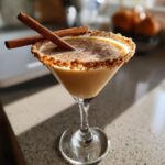 A festive Thanksgiving Pumpkin Pie Martini in a martini glass, rimmed with graham cracker crumbs and garnished with cinnamon sticks.