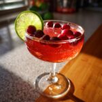 A festive Valentine’s Day Cupid’s Cranberry Kiss cocktail in a coupe glass, garnished with cranberries and a lime slice.