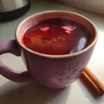 A close-up of a mug filled with Valentine’s Day Red Hot Cinnamon Cider, sprinkled with cinnamon and garnished with cinnamon sticks.