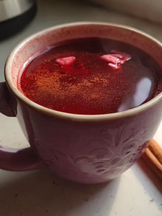 A close-up of a mug filled with Valentine’s Day Red Hot Cinnamon Cider, sprinkled with cinnamon and garnished with cinnamon sticks.