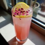 A tall glass of pink Valentine’s Day Rose Petal Lemonade topped with a lemon slice and dried rose petals.