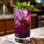A tall glass of Blueberry Mint Lemonade filled with ice, blueberries, mint leaves, and a vibrant purple hue.