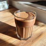 A glass of frothy Cacao Almond Energy Shake sits on a wooden surface, catching the sunlight.