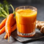 A refreshing glass of vibrant orange carrot ginger juice, with whole carrots and ginger root in the background.