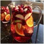 A pitcher filled with Cranberry Orange Mock Sangria, featuring cranberries, orange slices, apple slices, and ice.