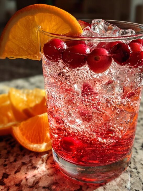 A glass filled with ice, cranberries, and a vibrant red liquid, garnished with an orange slice. This is the Cranberry Orange Vodka Twist.