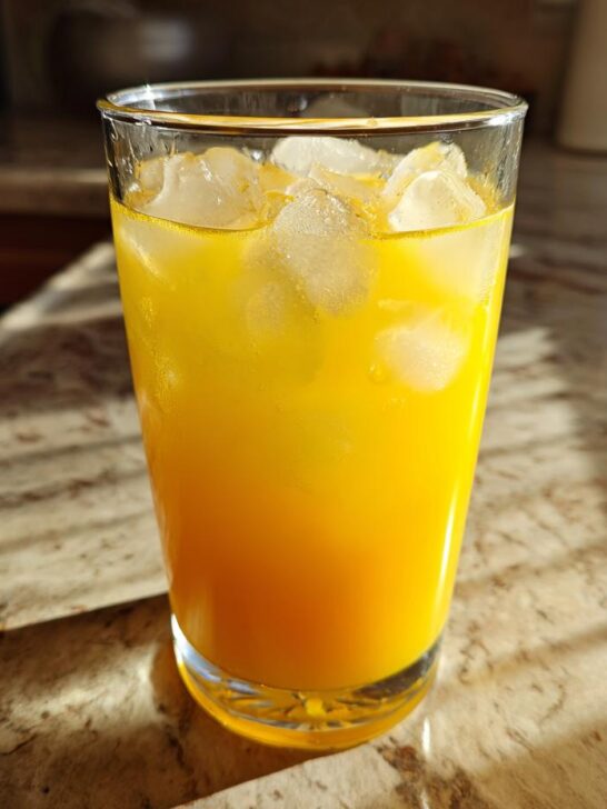 A tall glass filled with a vibrant yellow Honey Lemon Turmeric Spritzer and ice cubes, catching the sunlight.