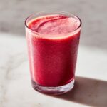 A tall glass filled with a vibrant red Kale Kiwi Energy Refresher drink, topped with foam.
