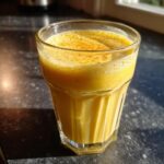 A vibrant yellow Mango Turmeric Recovery Drink in a glass, topped with foam and a sprinkle of spice.