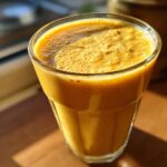A vibrant orange Mango Turmeric Recovery Drink in a tall glass, with a foamy top, bathed in sunlight.