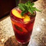 A tall glass filled with a vibrant red Peach Hibiscus Cooler, garnished with peach slices and fresh mint leaves.
