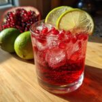 A close-up of a Pomegranate Lime Refresher in a glass with ice, garnished with lime slices and pomegranate seeds.