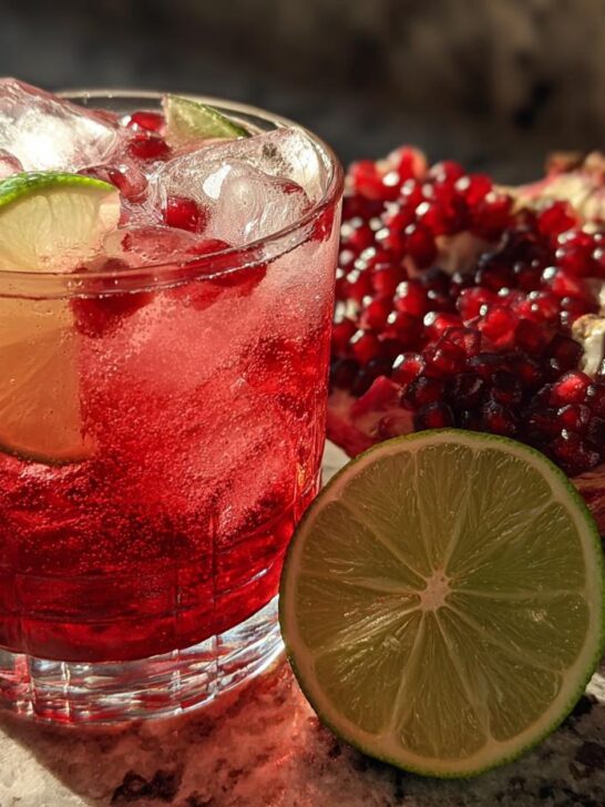 A vibrant Pomegranate Lime Refresher in a glass with ice, lime slices, and pomegranate seeds, next to a halved pomegranate.