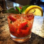 A close-up of a glass filled with Strawberry Basil Lemonade, featuring fresh strawberries, basil leaves, ice, and a lemon slice garnish.