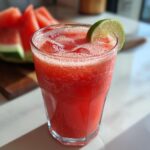 A tall glass filled with a vibrant pink Watermelon Ginger Refresher, topped with ice and garnished with a lime slice. Sliced watermelon is visible in the background.