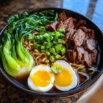 A close-up of a steaming bowl of beef ramen, featuring tender beef, ramen noodles, bok choy, and a perfectly halved soft-boiled egg.