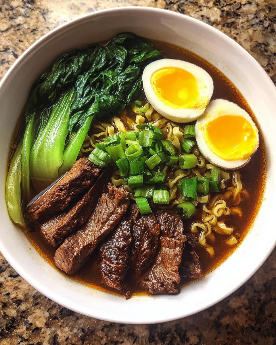 A close-up overhead shot of a bowl of Beef Ramen Recipe, featuring tender beef slices, ramen noodles, bok choy, a soft-boiled egg, and green onions.