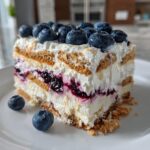 A perfect slice of Blueberry Cheesecake Icebox Cake showing layers of cream, graham crackers, and blueberry filling, topped with fresh blueberries.