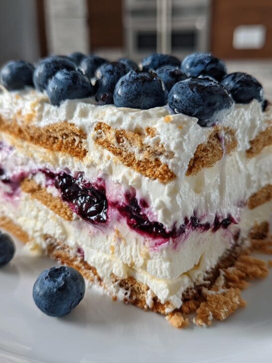 A perfect slice of Blueberry Cheesecake Icebox Cake showing layers of cream, graham crackers, and blueberry filling, topped with fresh blueberries.