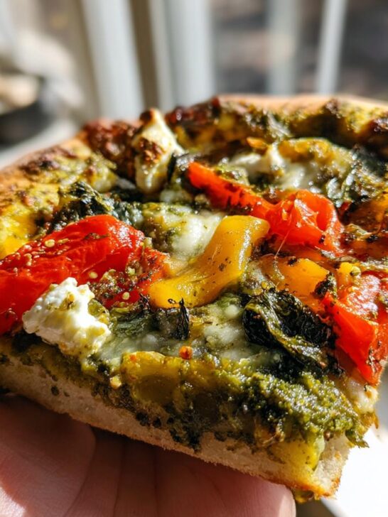 A slice of Chickpea Veggie Gluten Free Pesto Pizza held in a hand, showing pesto, vegetables, and cheese.