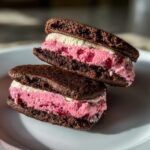 Two rich Chocolate Raspberry Sandwich Cookies stacked on a white plate, showing dark chocolate wafers and thick pink and white filling.