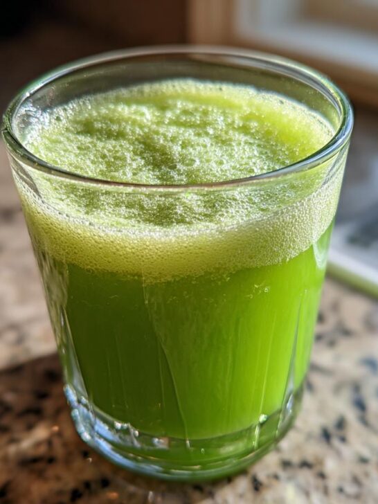 A close-up of a glass filled with vibrant green Cucumber Ginger Lemon Detox Juice, topped with foam.