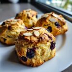 Close-up of four golden-brown Dairy Free Blueberry Almond Scones topped with sliced almonds on a white plate.