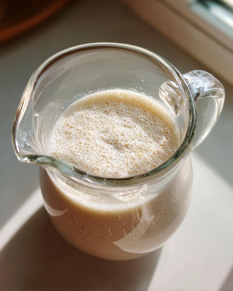 A glass pitcher filled with frothy, creamy Easy Homemade Oat Milk, catching the sunlight.