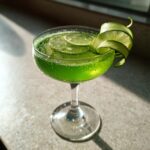 A vibrant green El Pepino cocktail in a coupe glass, garnished with a cucumber ribbon and lime slices.