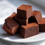 Close-up of rich, dark chocolate cubes of Fantasy Fudge dusted heavily with cocoa powder, piled on a white plate.