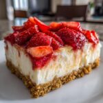 A close-up of a thick slice of Fresh Strawberry Cheesecake Bars with a graham cracker crust, topped with glazed strawberries.