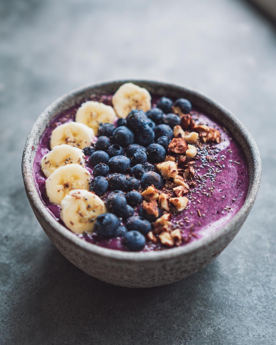 A vibrant purple smoothie bowl topped with sliced bananas, blueberries, chopped nuts, and chia seeds. A perfect guilt-free treat from our 10 Irresistible Frozen Banana Recipes.