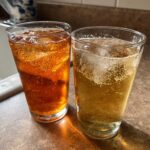 Two glasses filled with ice and bubbly beverages, one dark amber and the other lighter gold, representing Ginger Ale Vs Ginger Beer.