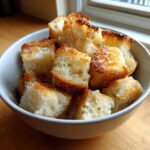 A close-up of a white bowl filled with golden brown, crispy homemade croutons.