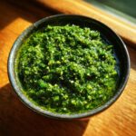 A close-up overhead view of vibrant green homemade pesto sauce in a dark bowl, showcasing its texture and fresh ingredients.