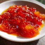A close-up of glossy, vibrant red Irresistible Sweet and Sour Sauce with visible seeds in a white bowl.