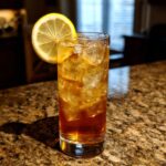 A tall glass filled with ice and a golden Kentucky Buck Cocktail, garnished with a lemon slice.